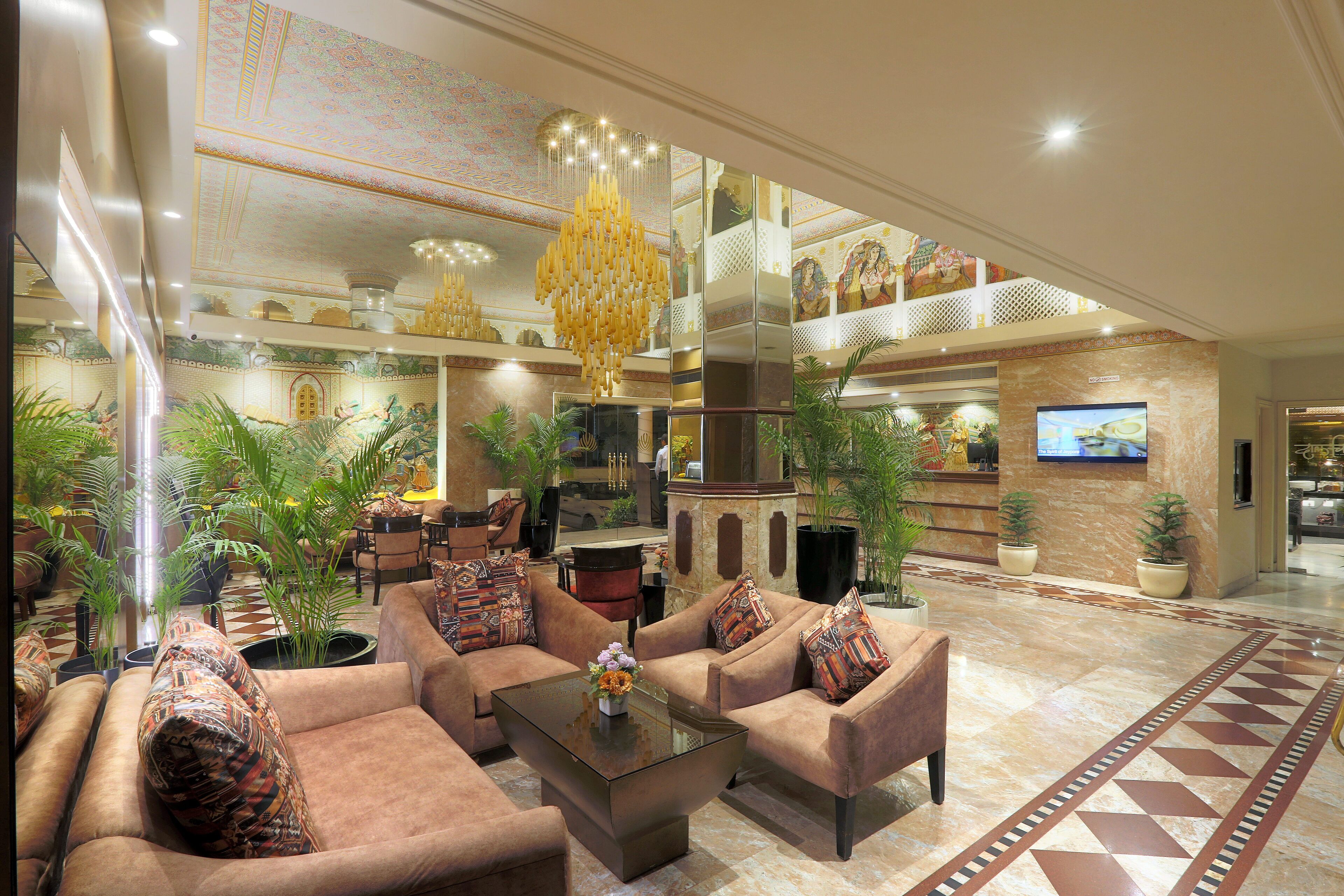 A spacious lobby with a high ceiling, a large chandelier, and a seating area with sofas and a coffee table. There are potted plants and a television mounted on the wall.
