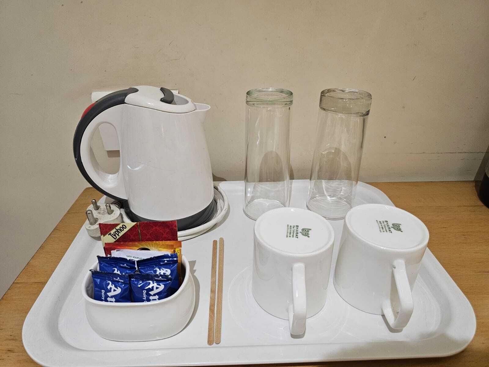 A tray with a white electric kettle, two glass tumblers, two white mugs, a small bowl with blue packets, and chopsticks.