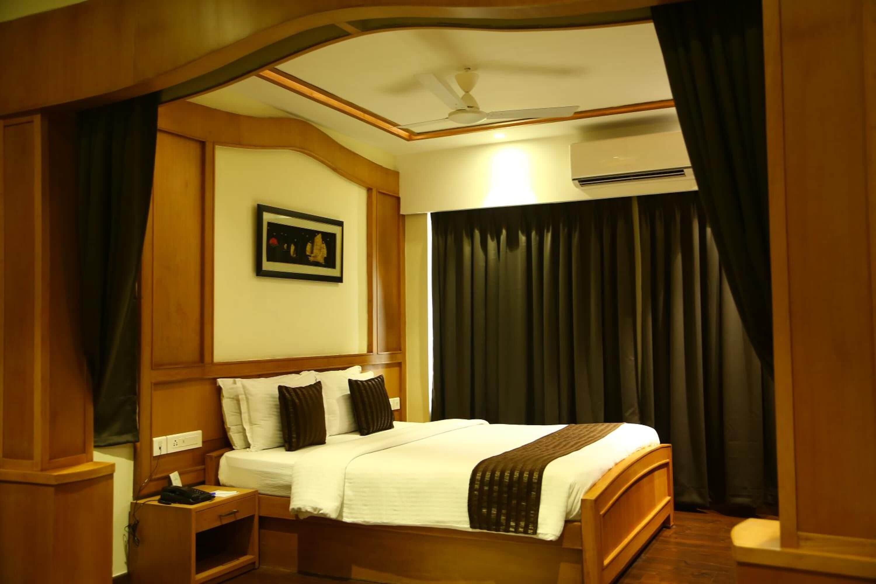 Una habitación de hotel con una cama de madera, una mesita de noche, un ventilador de techo y un ventanal grande con cortinas.
