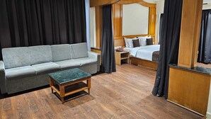 A hotel room with a grey sofa, a small wooden table, a bed with white bedding, and wooden paneling. - Hotel Mount Manor (Chennai)