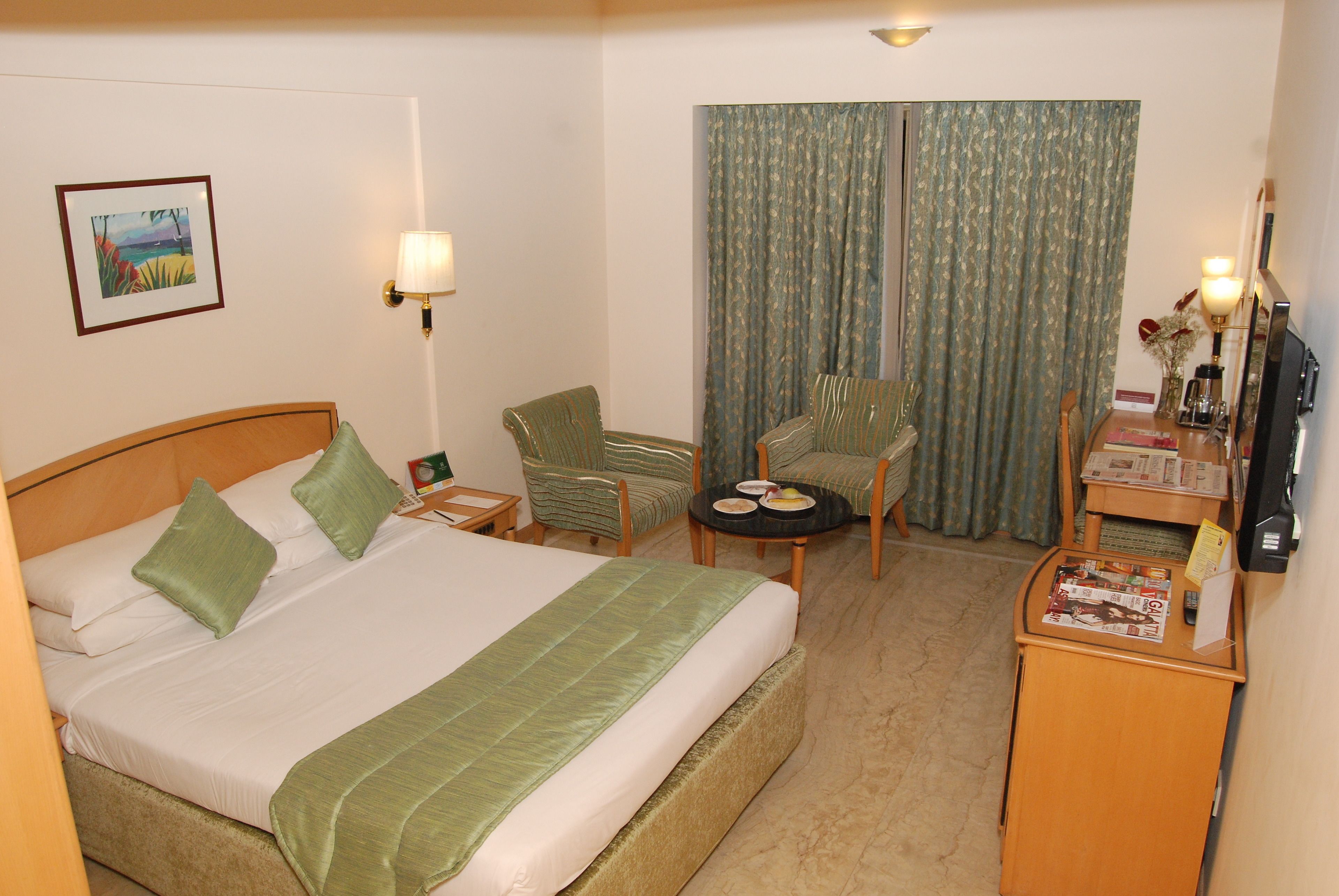 Habitación de hotel con cama, dos sillas, una mesita, un escritorio con computadora y una ventana con cortinas.