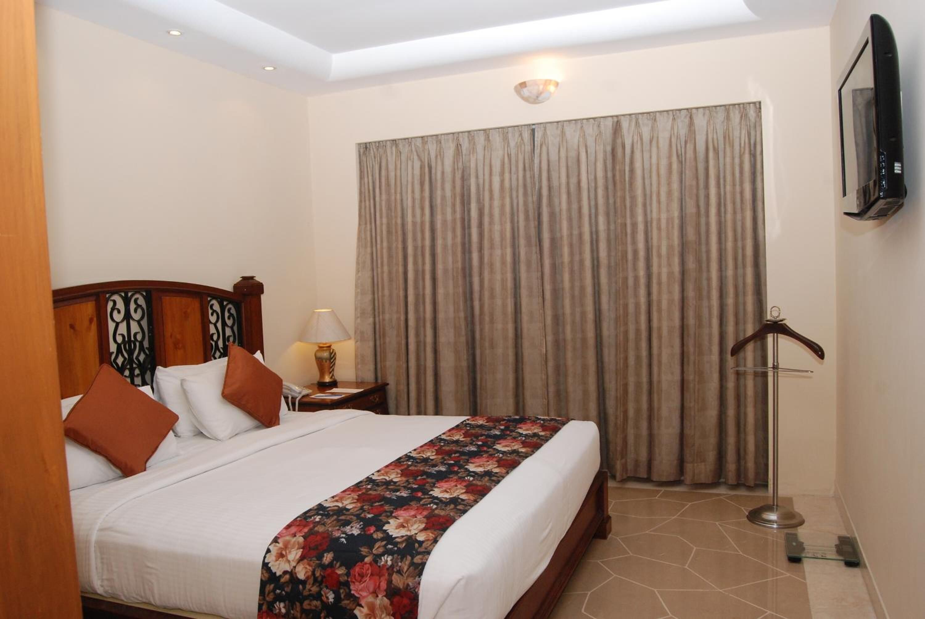 A hotel room with a bed, a nightstand, a lamp, a hanging coat rack, and a large window with curtains.