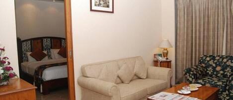 Une chambre d’hôtel avec un canapé beige, une table basse en bois, un fauteuil à motifs floraux, un lit avec une literie à motifs et un tableau encadré au mur.