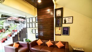 A reception area with a brown leather sofa, two armchairs, and a coffee table. There are framed awards and certificates on the wall, and a staircase with potted plants is visible in the background. - MY BIZZ HOTEL (Pune)
