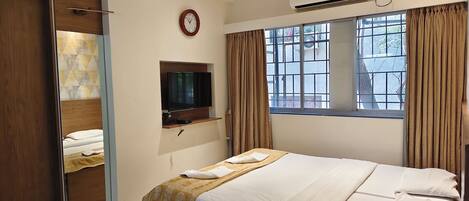 A hotel room with a bed, a TV mounted on a wooden shelf, a window with curtains, an air conditioning unit, and a clock on the wall.