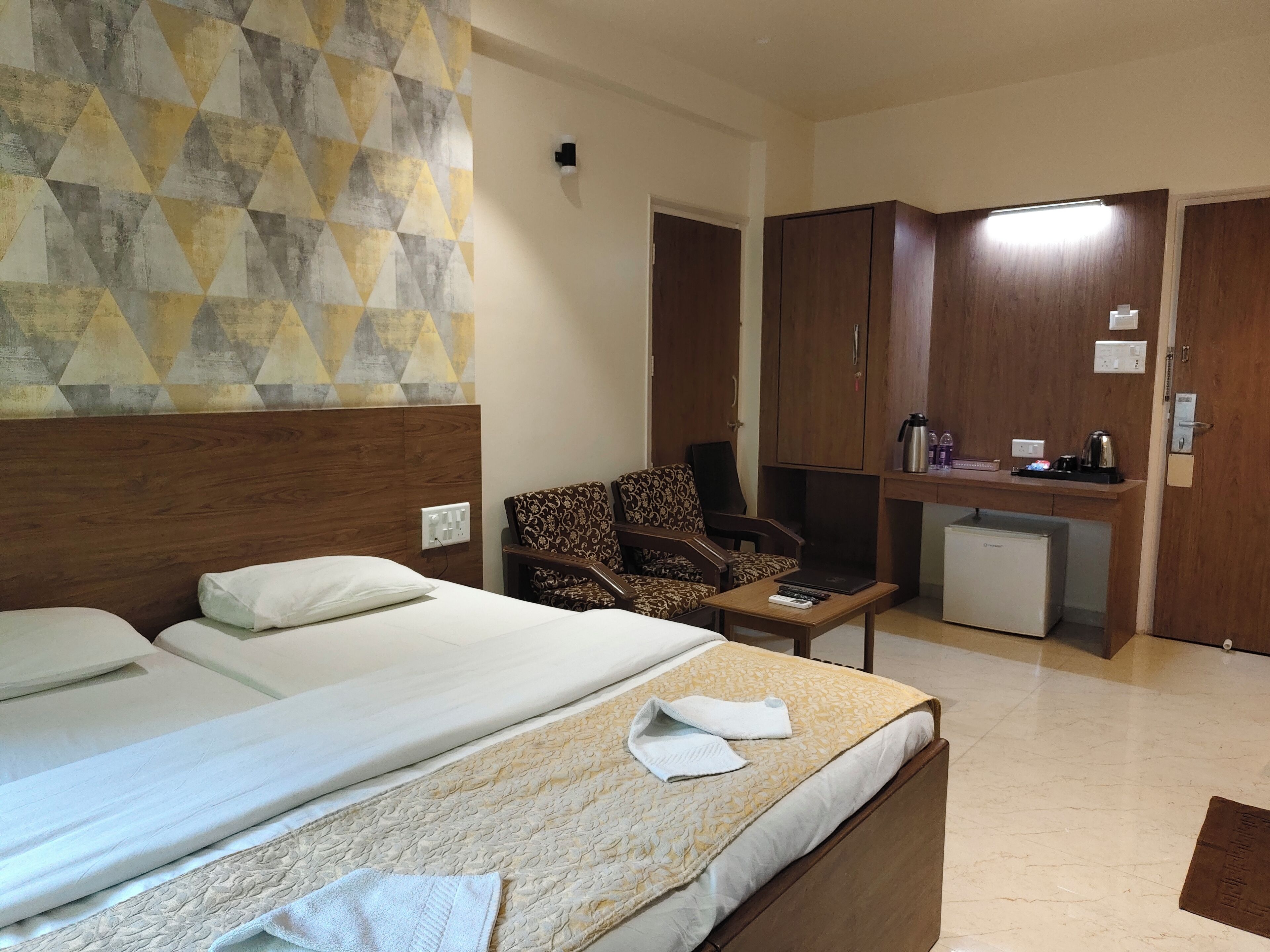A hotel room with two beds, a small desk, a chair, a kitchenette, and a wall with a geometric pattern.