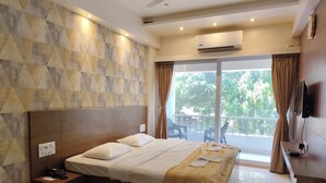 A hotel room with a bed, a desk with a laptop and remote controls, an air conditioner, and a view of greenery through the window. - Hotel Shreyas (Pune)