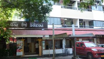 Ein Hotel namens 'Hotel Shereyas' mit einem mehrsprachigen Schild, ein rotes Fahrzeug vor der Fassade und ein Gebäude mit roten Ziegeldach.