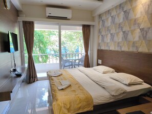 A hotel room with a bed, a TV mounted on the wall, an air conditioner, and a balcony with a table and chairs. - Hotel Shreyas (Pune)