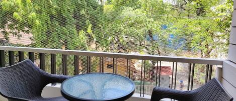 Un balcone con due poltrone in vimini e un tavolino in vetro, affacciato su un giardino con alberi e una recinzione.