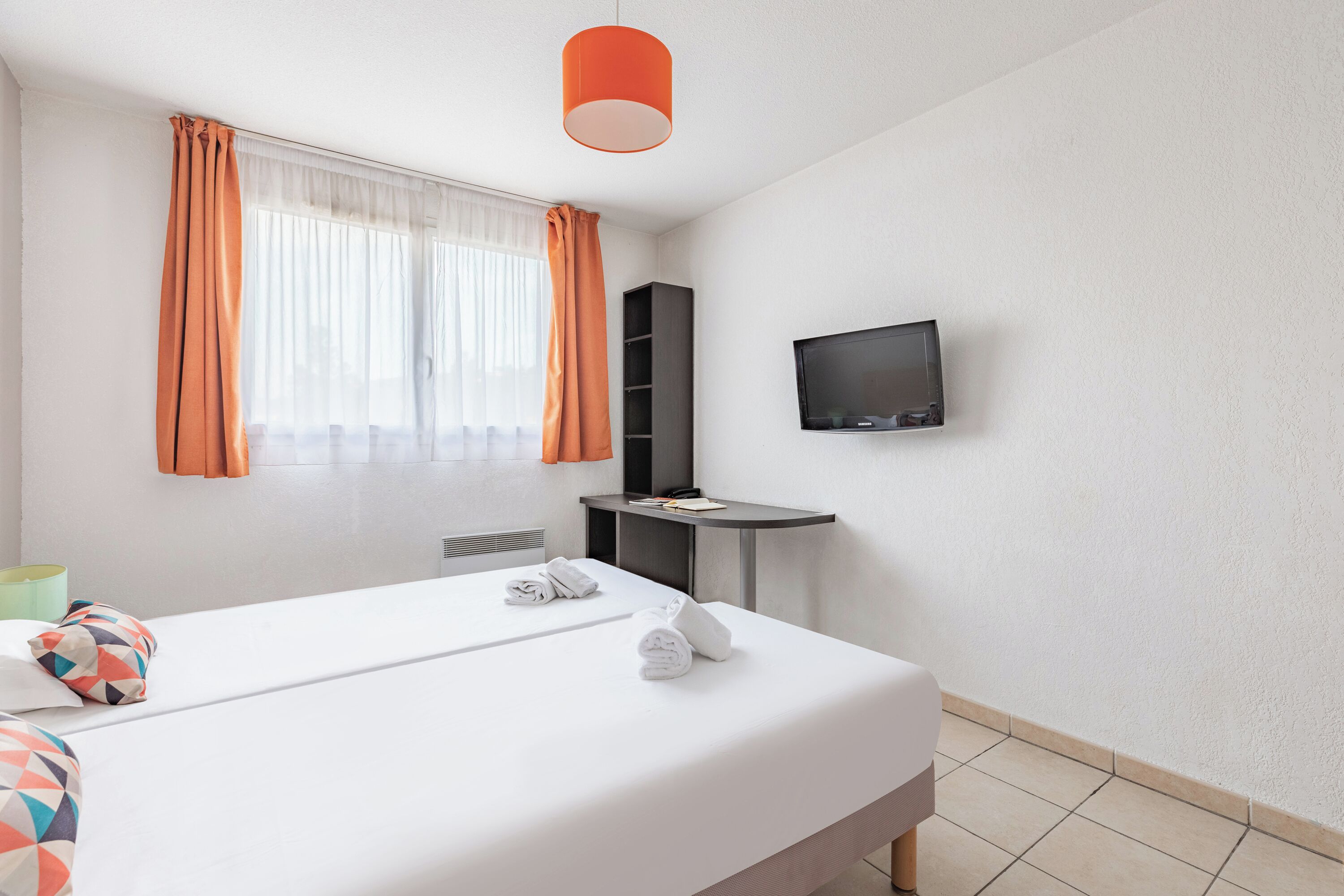 Une chambre d’hôtel avec un lit, un téléviseur fixé au mur, une petite table et une fenêtre avec des rideaux.