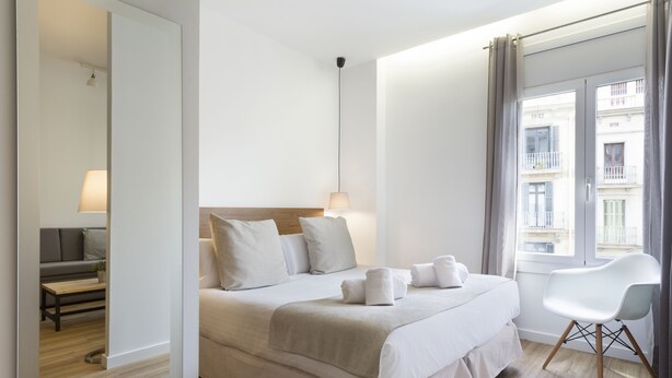 A modern hotel room with a large bed, a sitting area with a sofa and table, and a view of the city through the window. - Amister Apartments (Barcelona)