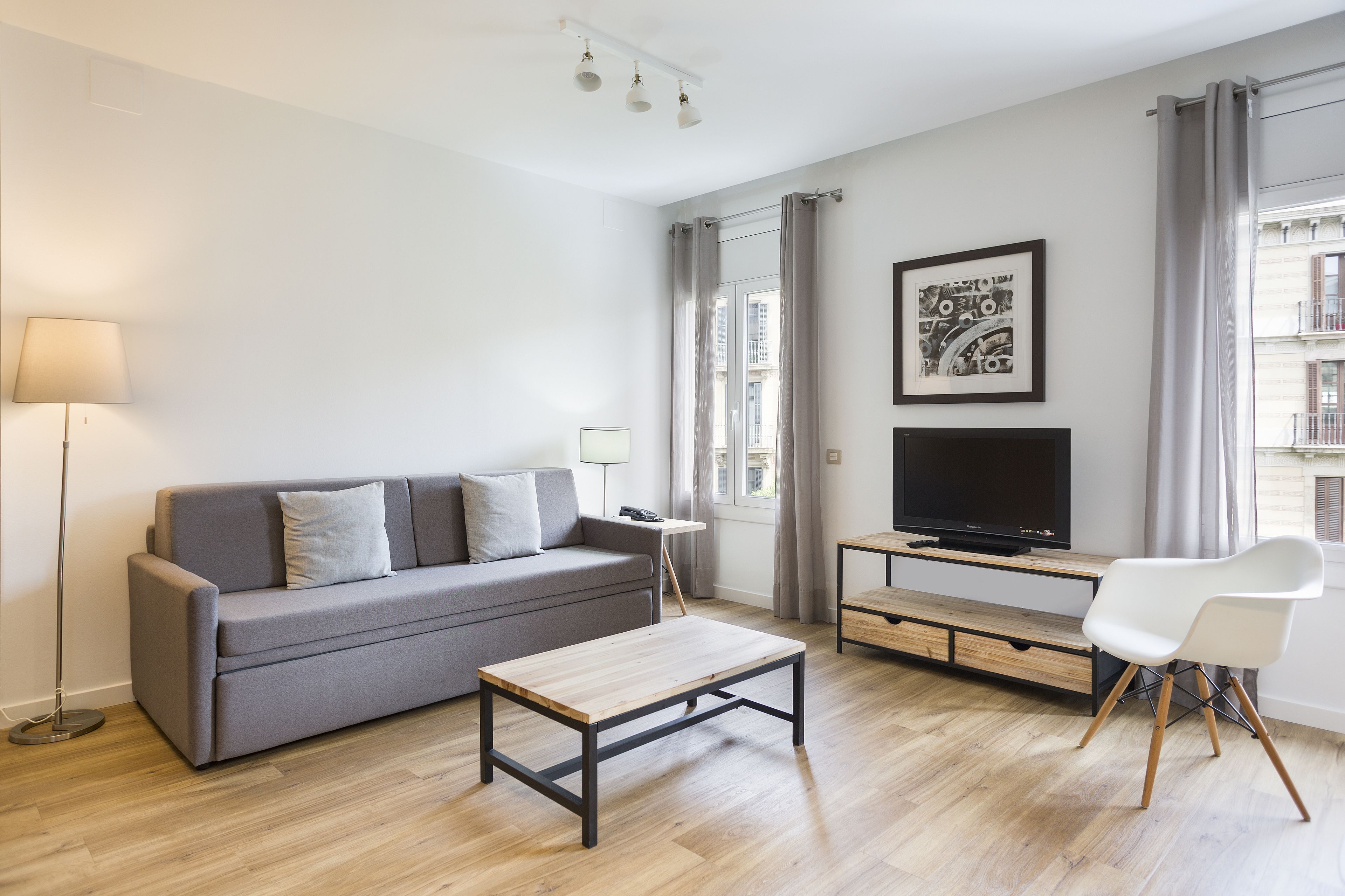 Una sala moderna con un sofá gris, una mesa de centro de madera, un televisor sobre un mueble de madera, una silla blanca y una lámpara de pie.