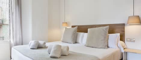 Una cama bien hecha con almohadas y toallas, una mesita de noche con lámpara, y una ventana con cortinas.