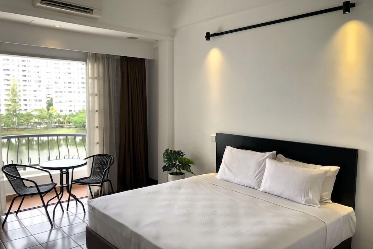 Une chambre d’hôtel avec un lit, une petite table avec des chaises, une plante en pot et une vue sur un immeuble par la fenêtre.