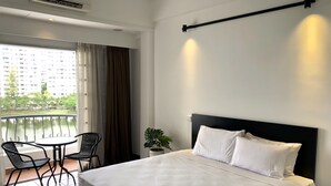 Une chambre d’hôtel avec un lit, une petite table avec des chaises, une plante en pot et un balcon donnant sur des immeubles et des arbres.