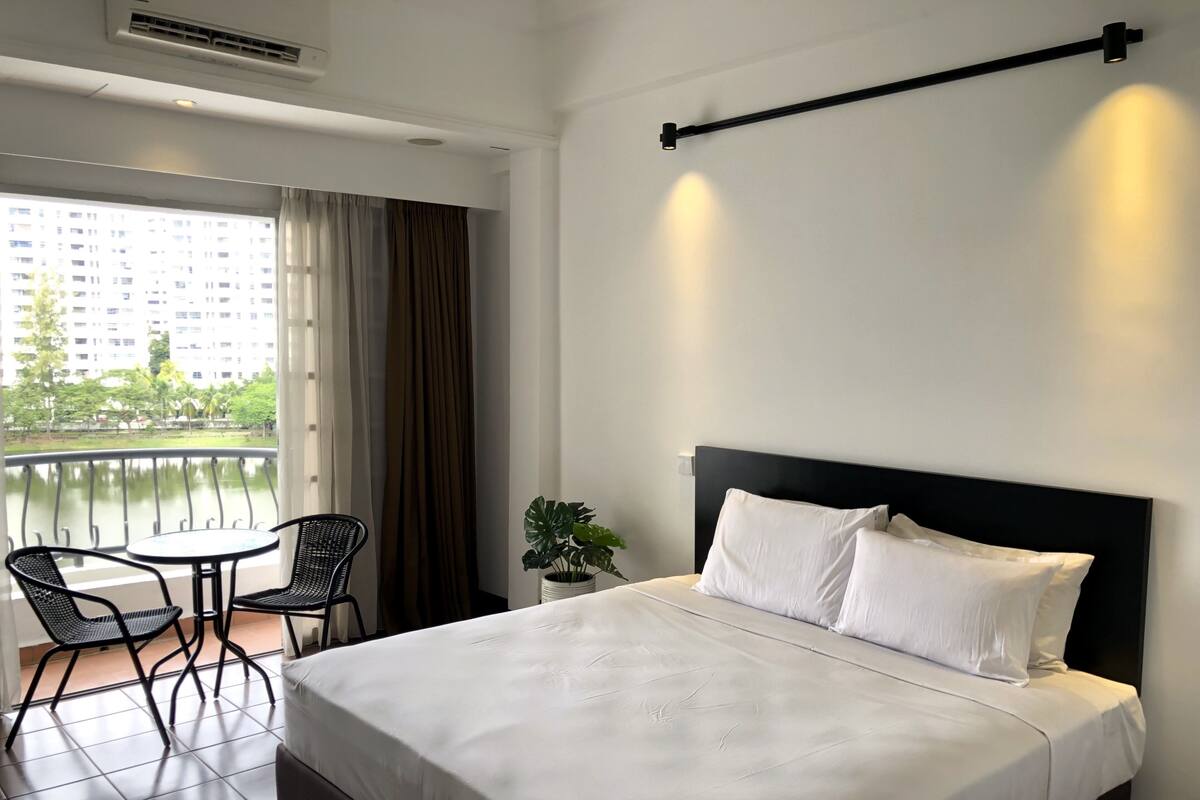 Une chambre d’hôtel avec un lit, une petite table avec des chaises, une plante en pot et un balcon donnant sur des immeubles et des arbres.