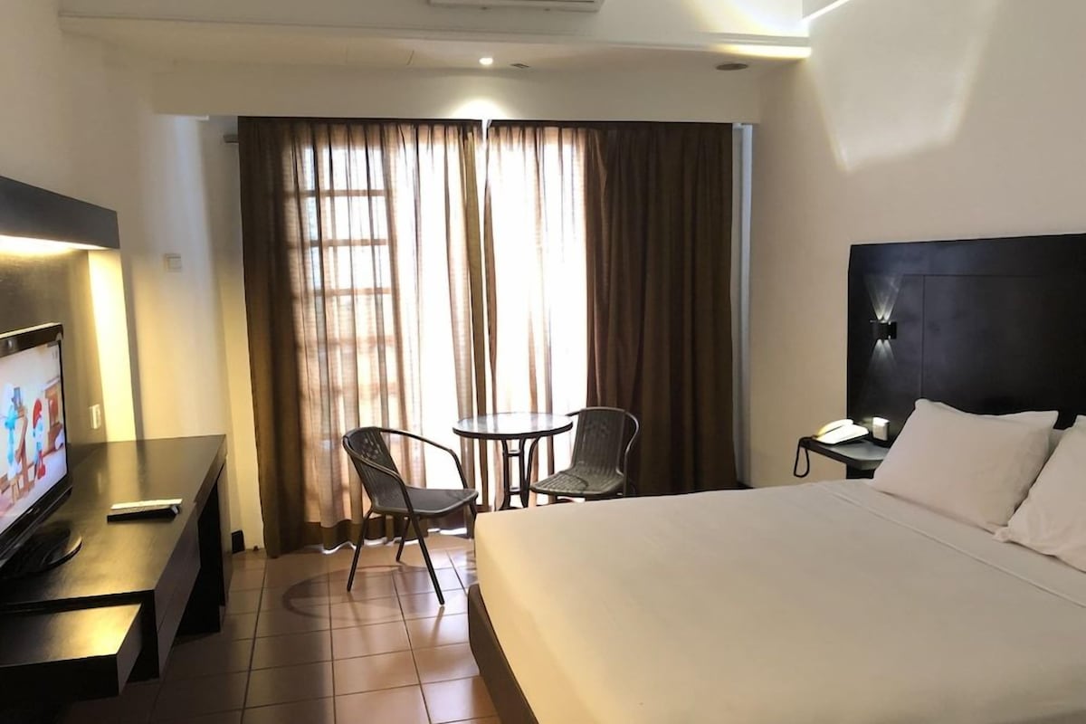 Une chambre d’hôtel comprenant un lit, un bureau avec une télévision, une chaise, une petite table et une fenêtre avec des rideaux.