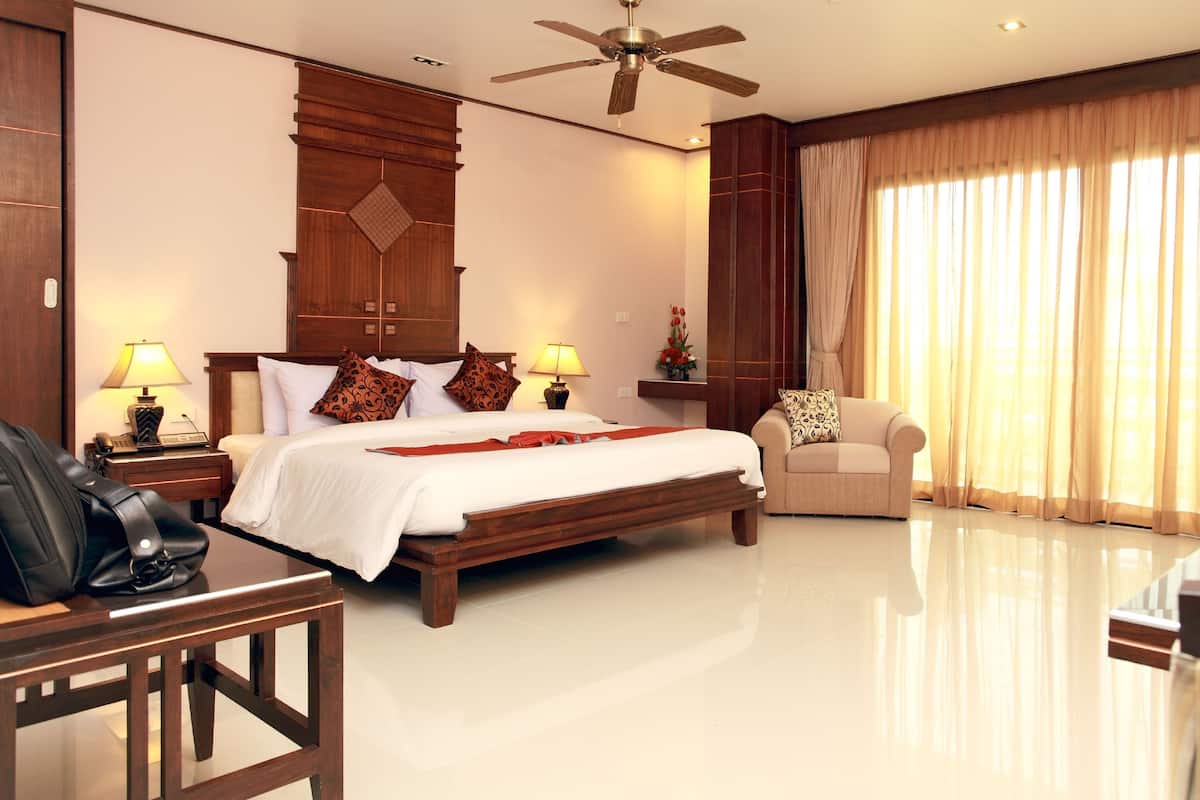 A hotel room with a large bed, a desk, a chair, a sofa, a ceiling fan, and a large window with curtains.