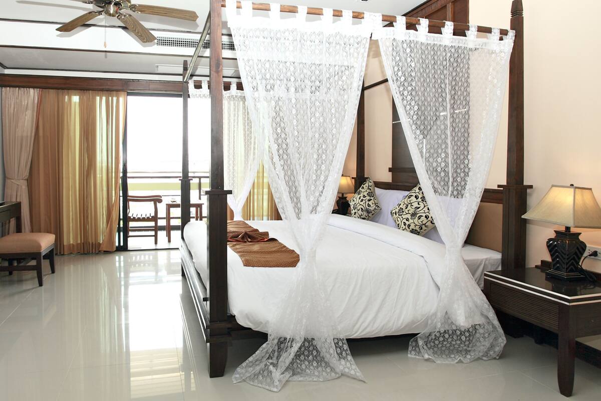 A four-poster bed with white bedding and lace canopy, a television on a wooden stand, a desk with a chair, and a balcony with curtains.