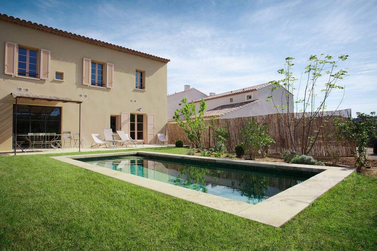Een huis met een zwembad, een terras met stoelen en een verzorgd gazon.