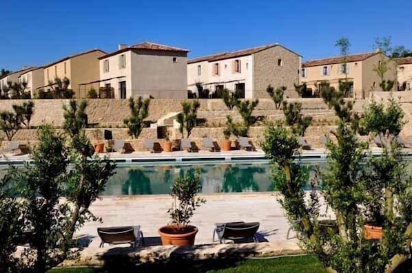 A poolside area with lounge chairs and potted plants, surrounded by stone buildings with terracotta roofs.