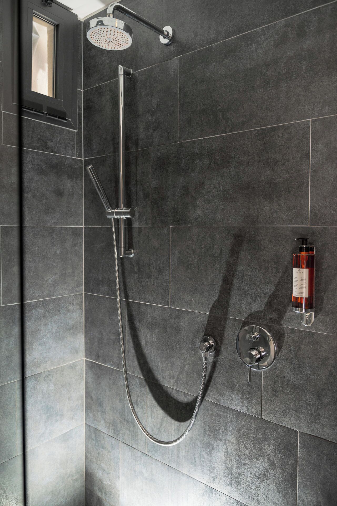 Una ducha moderna con cabezal de ducha de mano y ducha de lluvia, montada en una pared con azulejos oscuros.