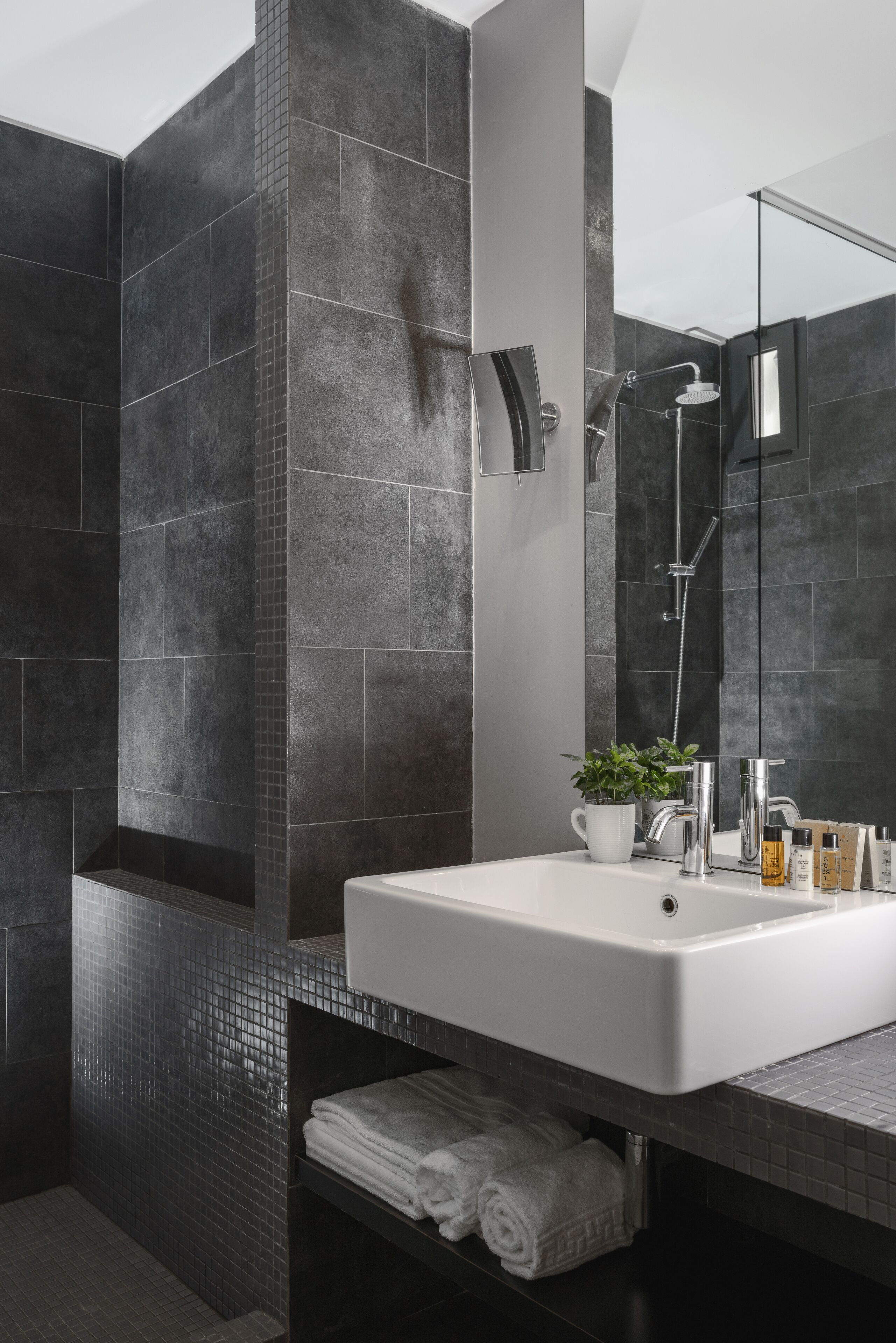 Un baño moderno con azulejos oscuros, un espejo grande, un lavabo blanco y una zona de ducha.