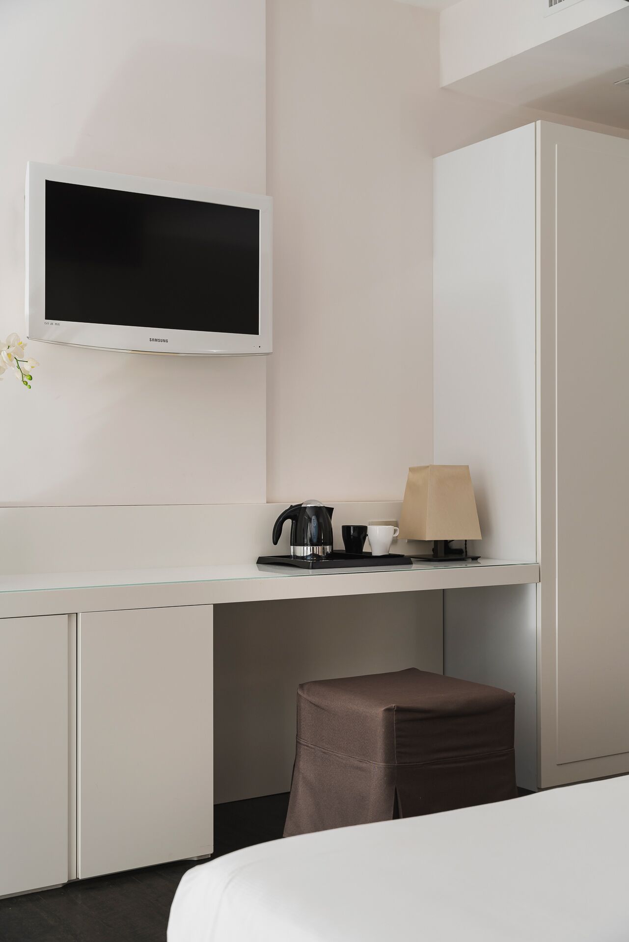 A hotel room with a bed, a desk with a television, a kettle, a cup, and a lamp.