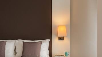 Una cama bien hecha con ropa de cama blanca y marrón, una lámpara de noche y una mesita de noche con un libro y una taza verde azulada.