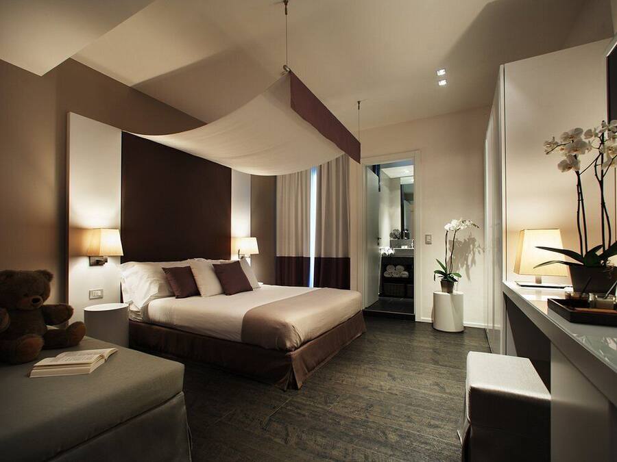 A modern hotel room with a large bed, bedside lamps, a desk, a chair, a teddy bear, and a potted plant.