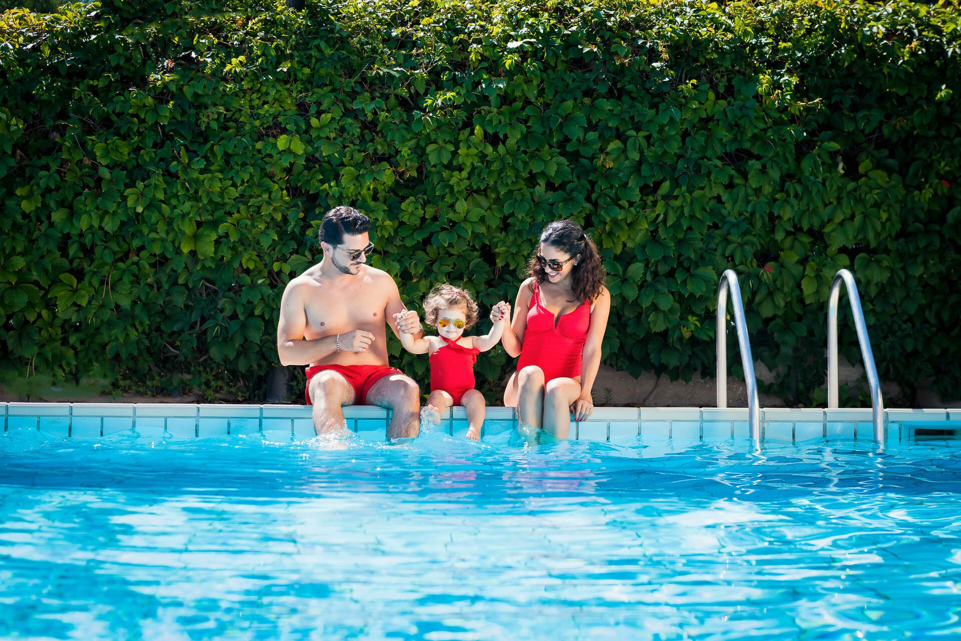 Una familia de tres junto a una piscina con un seto al fondo.