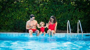 Una familia de tres junto a una piscina con un seto al fondo.