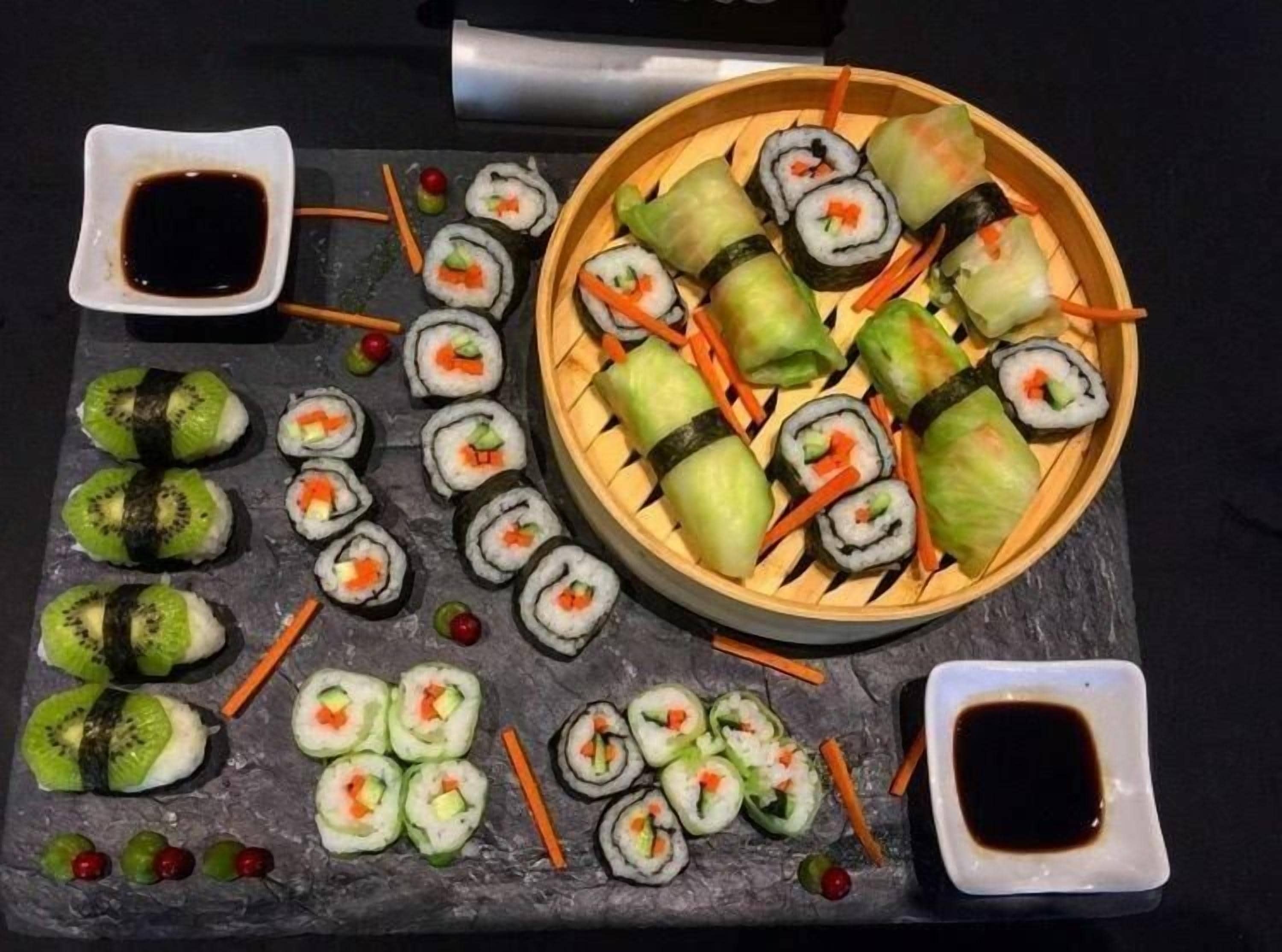 A variety of sushi rolls and nigiri on a slate board, accompanied by small bowls of soy sauce.