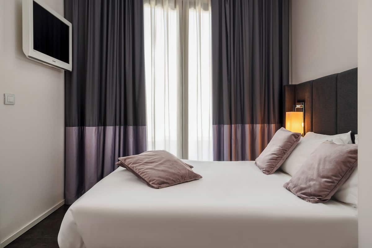 A hotel room with a bed, two pillows, a nightstand with a lamp, and a window with curtains.