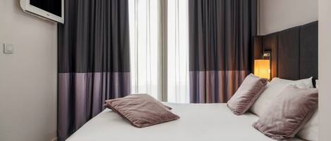 Habitación de hotel con cama, dos almohadas, mesita de noche con lámpara y ventana con cortinas.