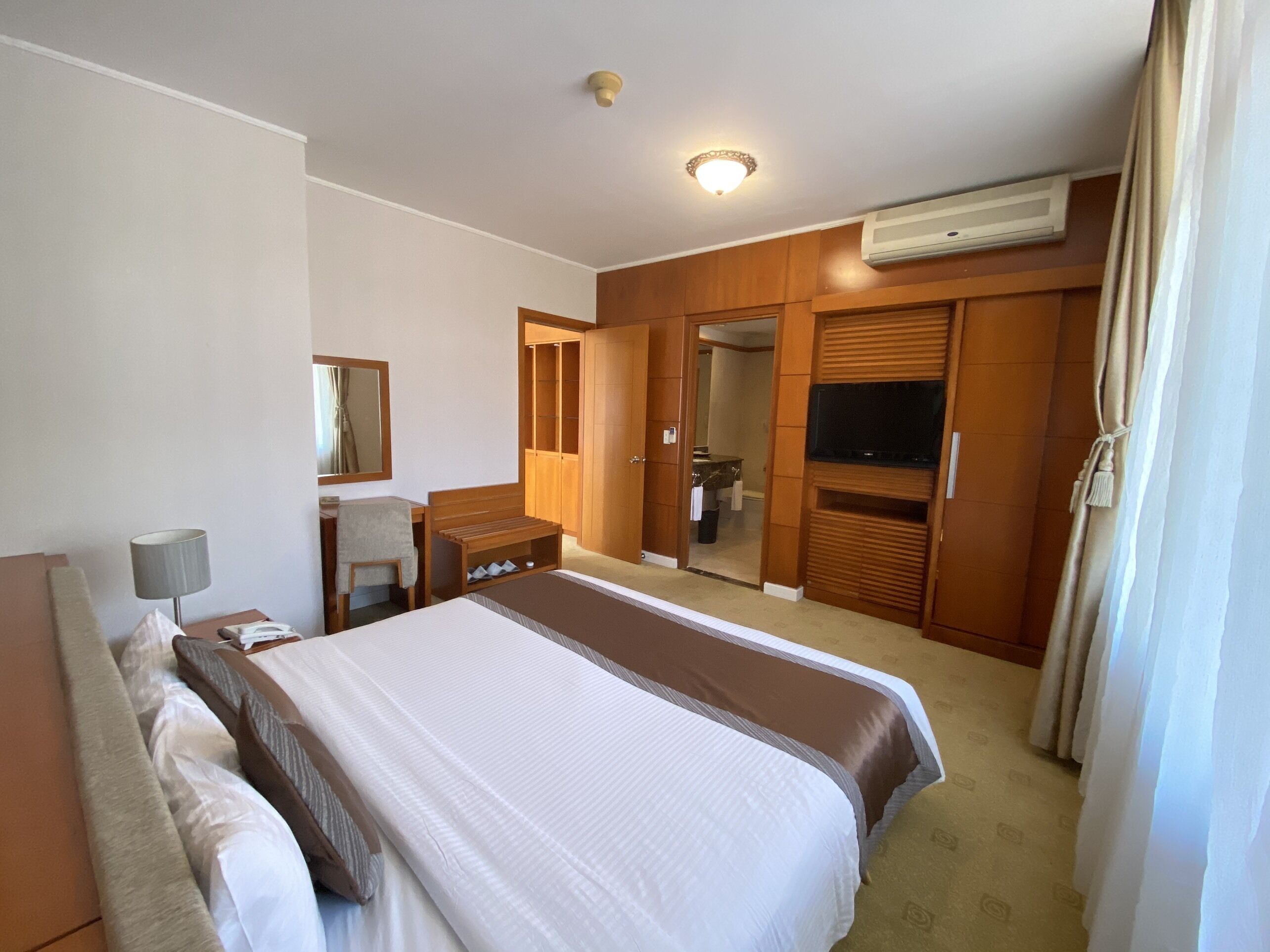 A hotel room with a large bed, a desk with a lamp, a TV, a wardrobe, and a bathroom visible through an open door.