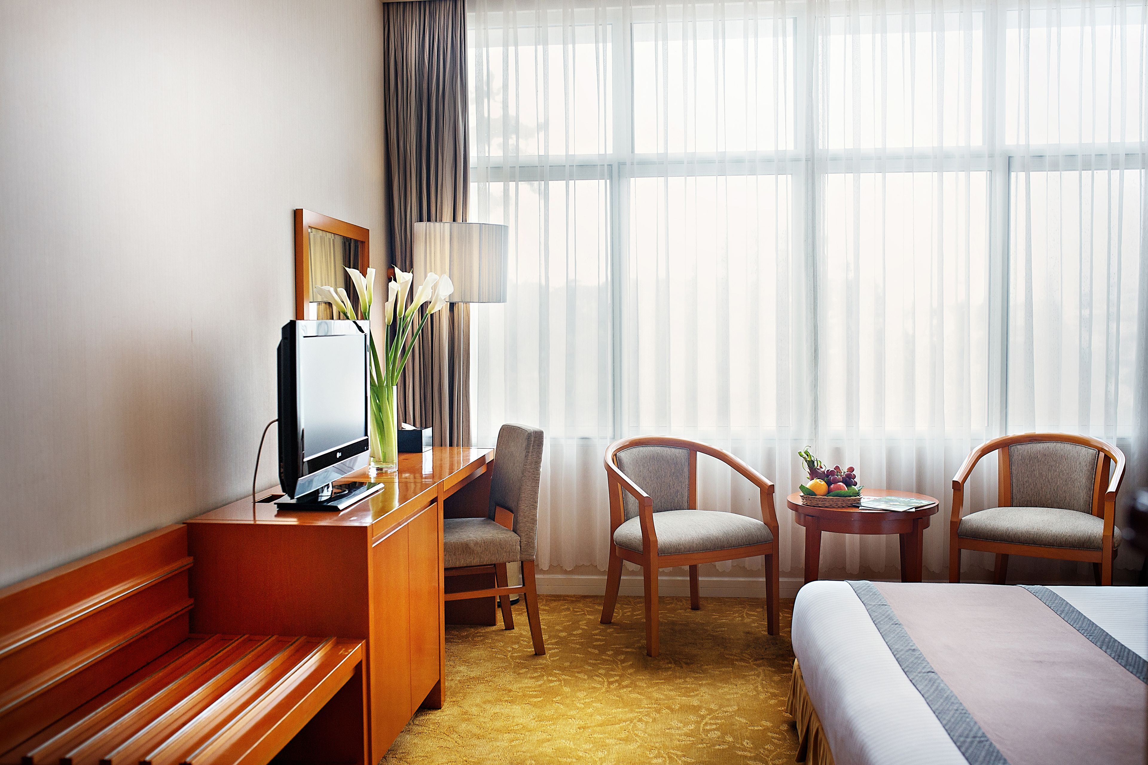 A hotel room with a bed, a desk with a television, two chairs, a small table with fruit, and a large window with sheer curtains.