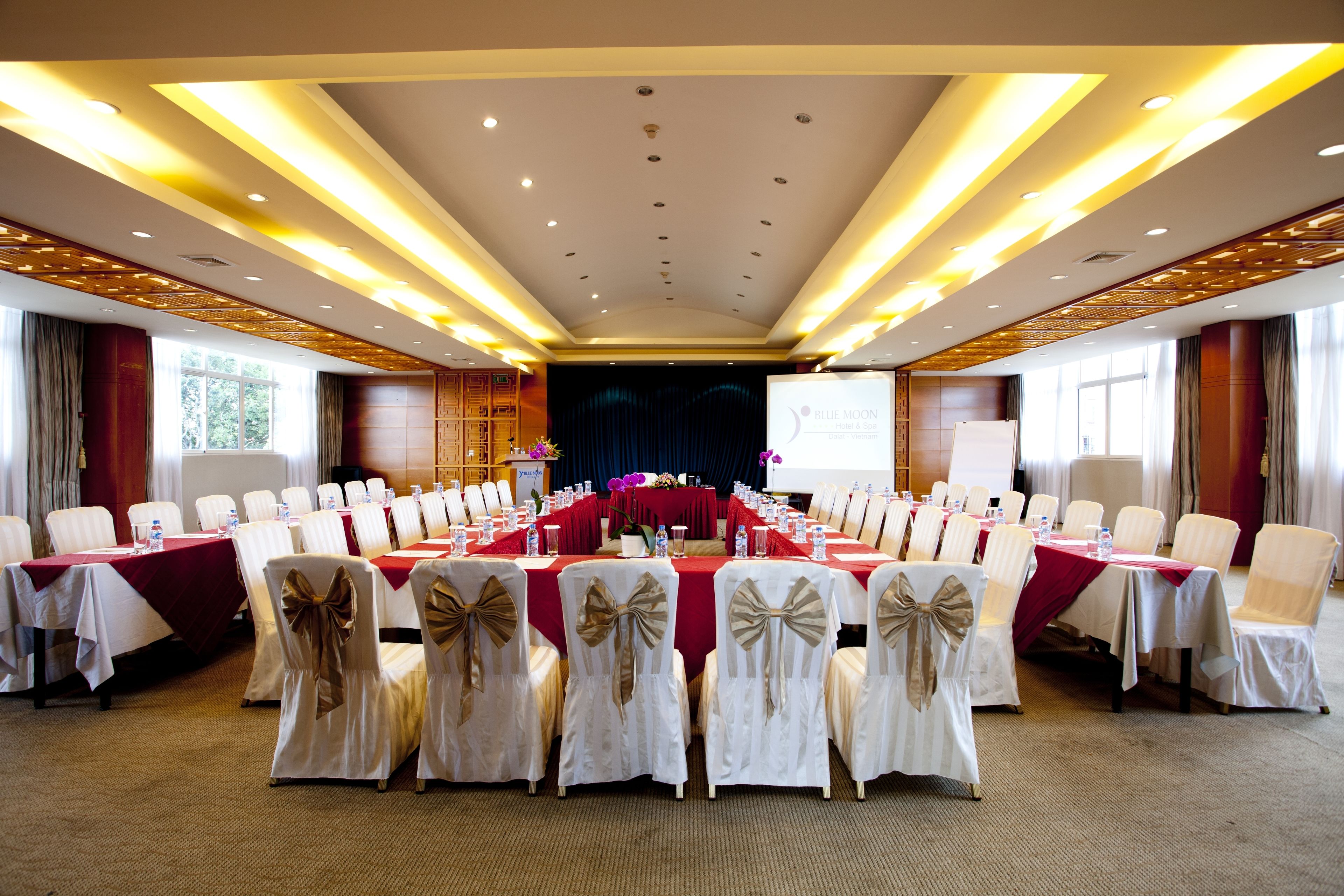 Une salle de conférence avec une longue table, des chaises blanches ornées de rubans dorés et une nappe rouge.