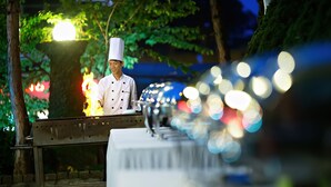 A chef cooking outdoors at night with a grill and a table in the foreground. - Muong Thanh Da Lat (Xuan Huong - Da Lat)