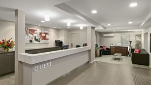 Eine moderne Hotelhalle mit einer Rezeption mit dem Schild "QUEST", einer Kaffeemaschine, einer Sitzecke mit einer Couch und Sesseln sowie einer dekorativen Wandgestaltung.