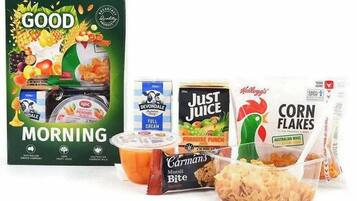 Uma mesa de café da manhã com Good Morning, iogurte integral Devondale, suco Paradise da Just Juice, Corn Flakes Kellogg's e Muesli Bite da Carman's.