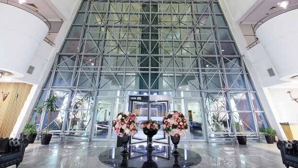 Un lobby moderno con fachada de vidrio, una mesa redonda central con arreglos florales y un techo alto con diseño geométrico.