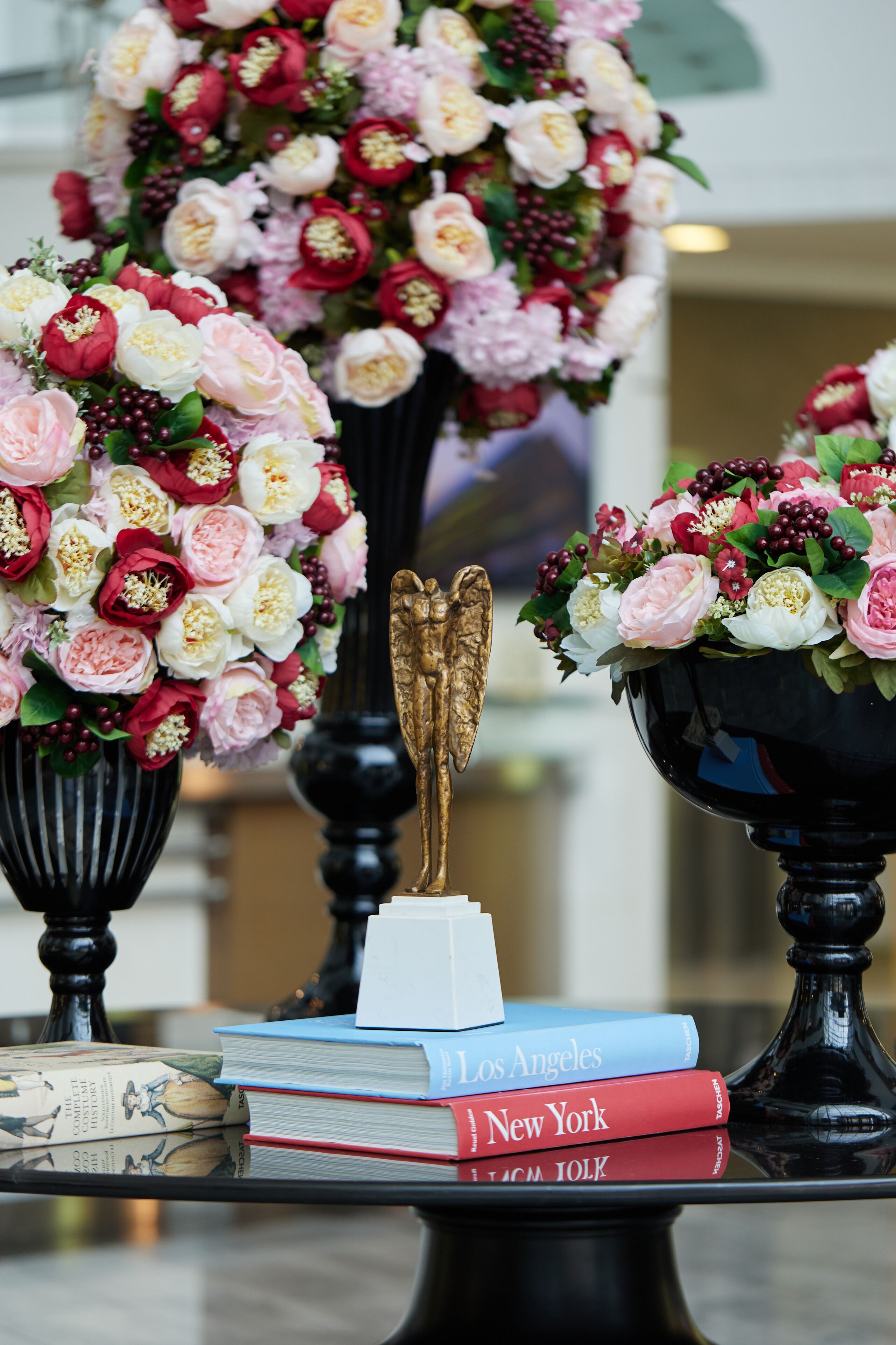 A table with two vases of flowers, a golden angel statue, and a stack of books titled 'Los Angeles' and 'New York'.