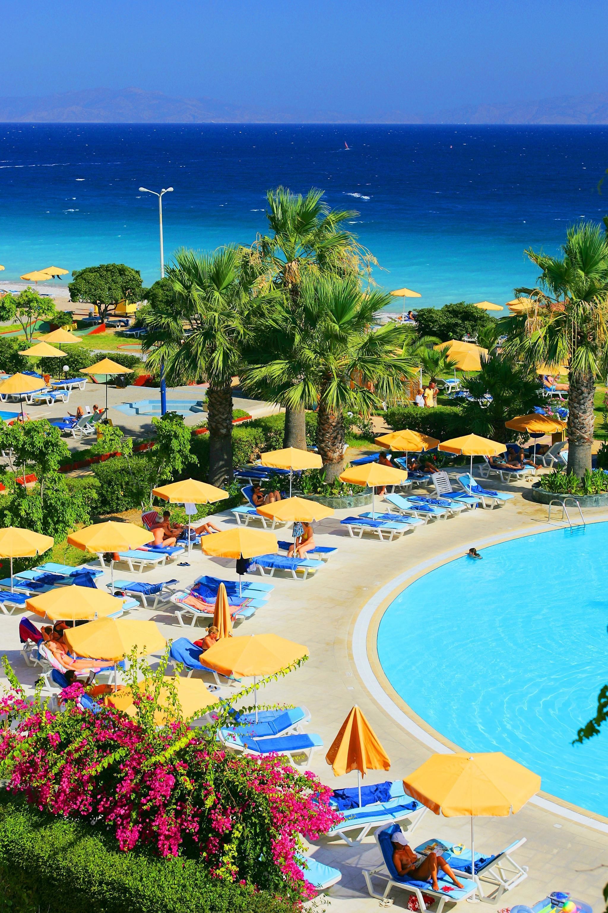 Un espace piscine aménagé dans un complexe hôtelier, avec des chaises longues et des parasols, entouré de palmiers et offrant une vue sur l’océan.