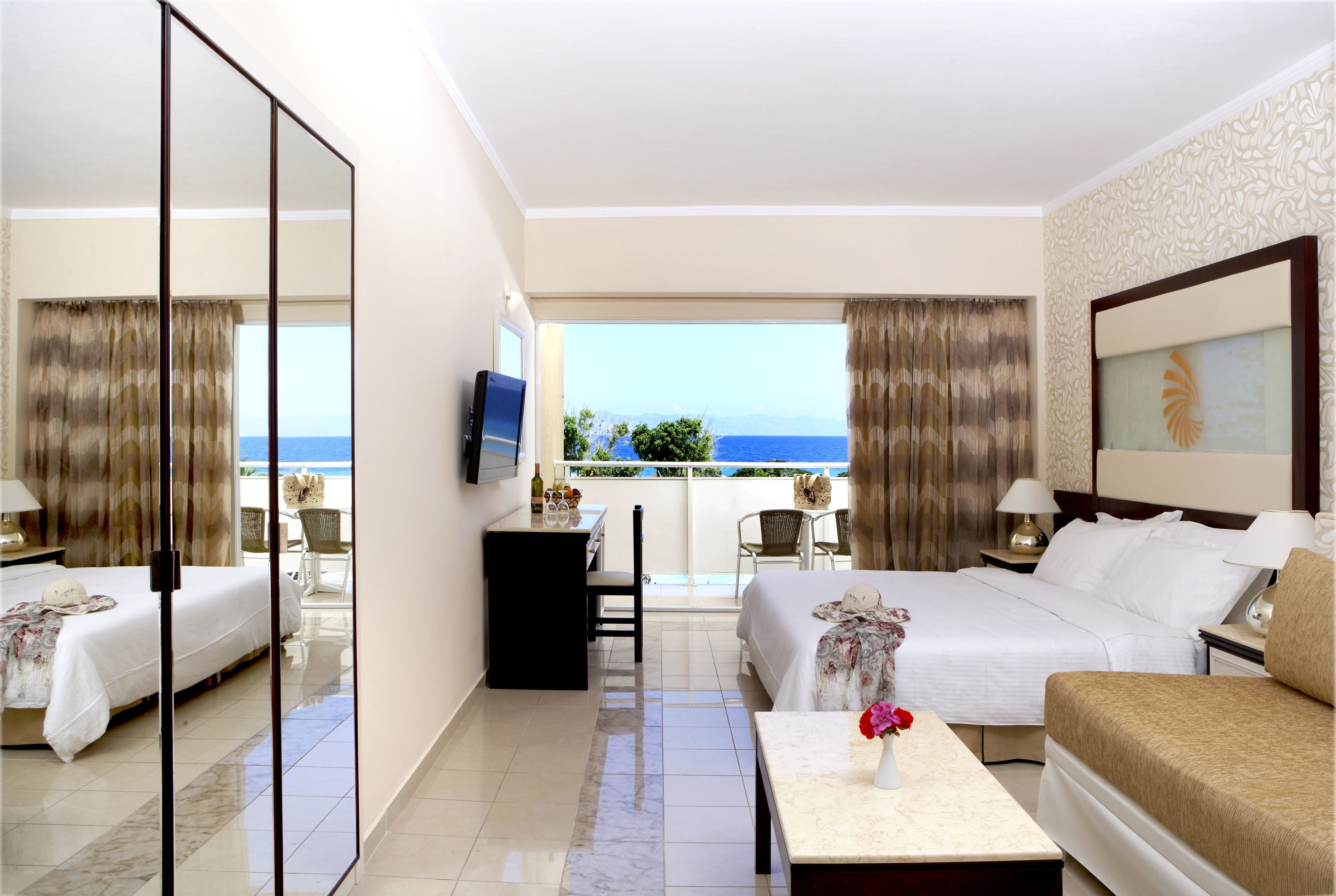 A hotel room with a bed, a desk, a chair, a TV, and a balcony with a view of the sea.
