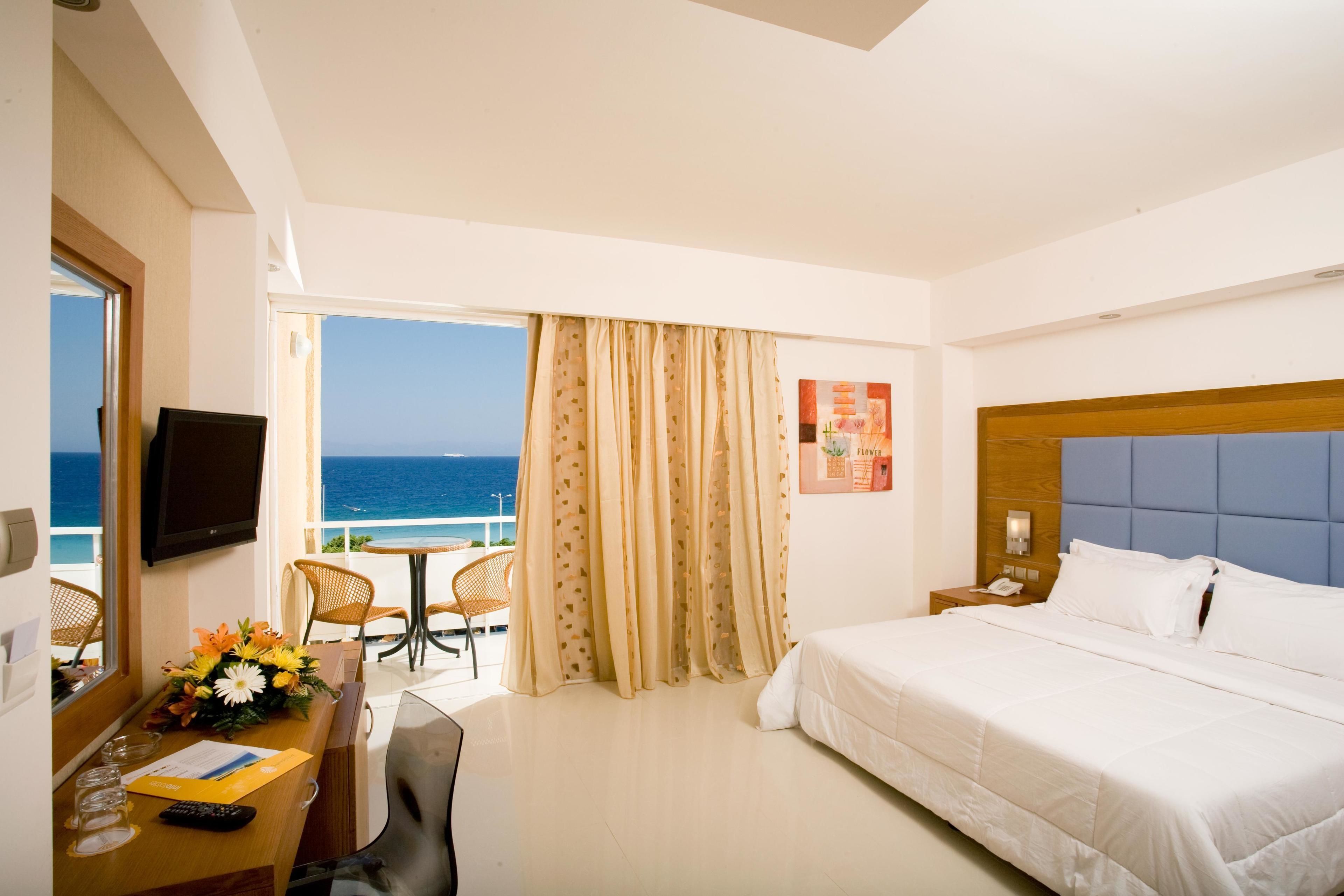 A hotel room with a bed, a desk with a computer, a chair, a table with flowers, a TV, and a view of the ocean.