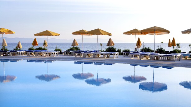 A poolside area with lounge chairs and umbrellas, a clear view of the sea, and a calm pool reflecting the surroundings.