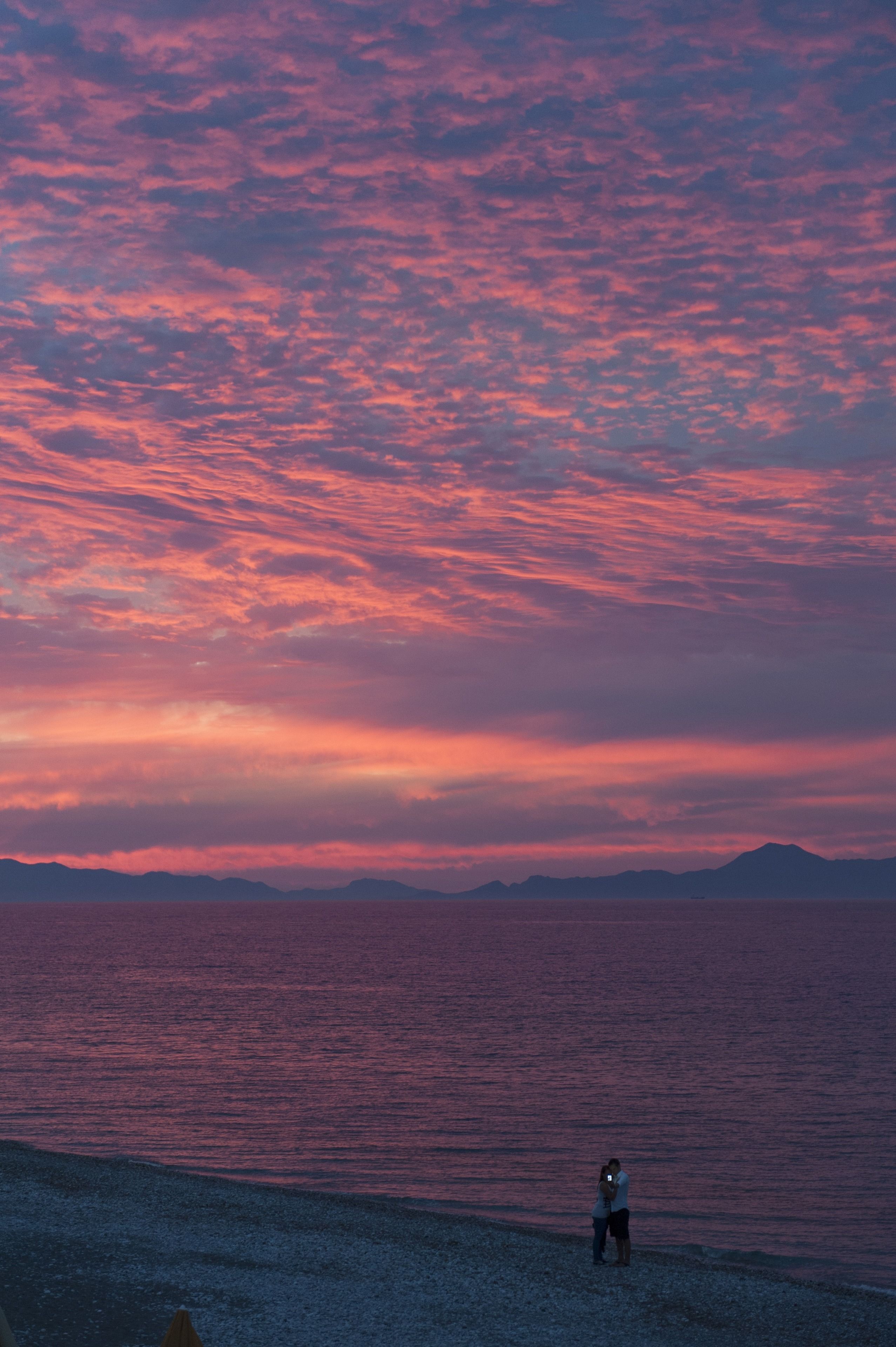A person standing on a pebbled beach with a view of the ocean and distant mountains under a pink and purple sky.