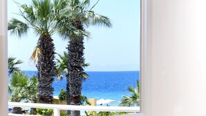 A window with a view of the ocean and palm trees, a bottle of wine, and a fruit bowl on a counter.
